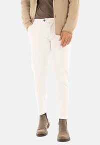 Man in witte slim-fit broek, bruin suède Chelsea boots, een bruin shirt en een beige textuur blazer met zijn handen in de zakken.