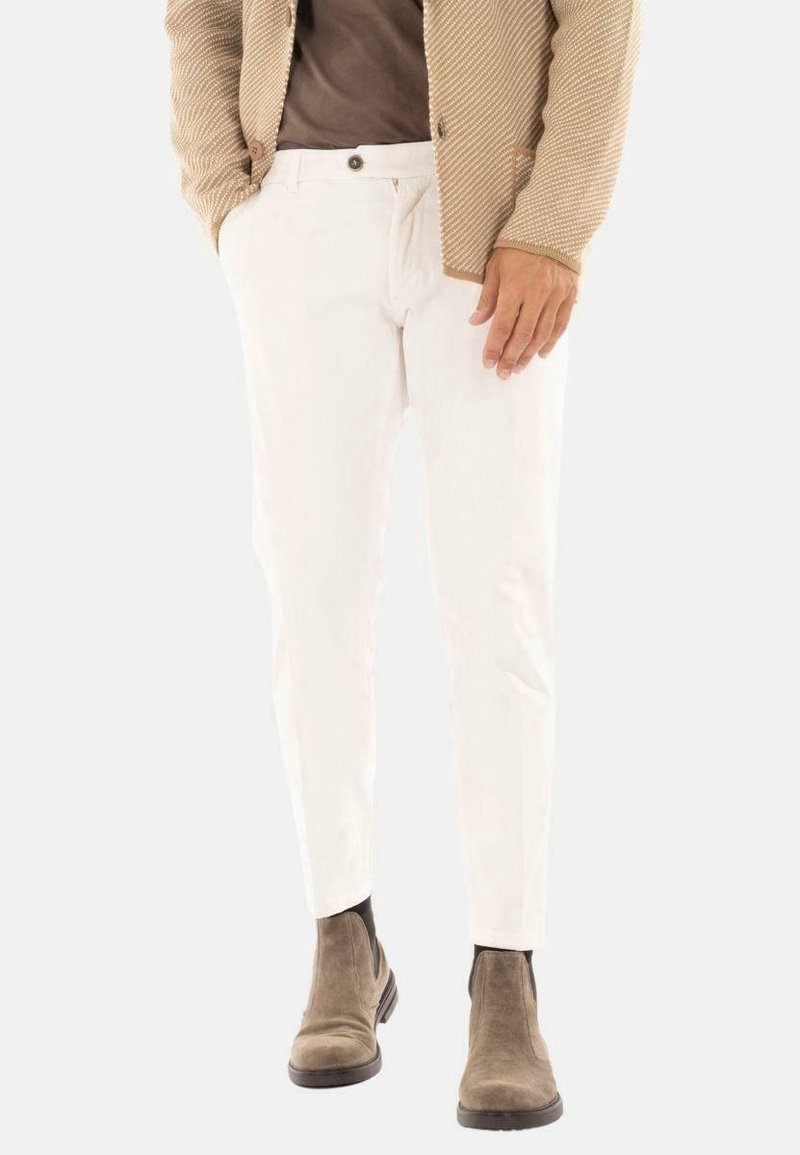Man in witte slim-fit broek, bruin suède Chelsea boots, een bruin shirt en een beige textuur blazer met zijn handen in de zakken.