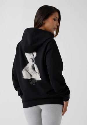 Donna che indossa una felpa nera con cappuccio con grafica di orsetto bianco e testo "Guess 1981" sulla schiena, abbinata a pantaloni grigio chiaro.