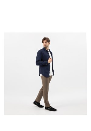 Uomo che cammina al chiuso indossando una camicia blu navy, t-shirt bianca, pantaloni beige e scarpe slip-on nere su uno sfondo bianco uniforme.