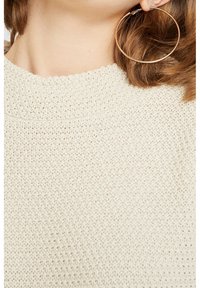 Pull en maille crème avec un motif texturé, doté d'un col haut. Grande boucle d'oreille en or apporte un accent métallique contrastant.