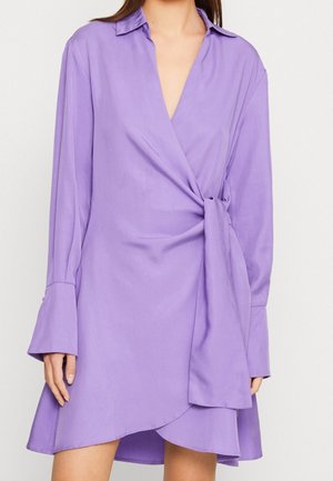 Kvinde iført en langærmet lilla slå-om-kjole med krave og knyttedetalje i taljen.