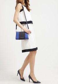 Bílé šaty s námornickými horizontálními pruhy, doplněné modro-černou kabelkou přes rameno. Modelka má na sobě námornické vysoké podpatky. Hladká textura.