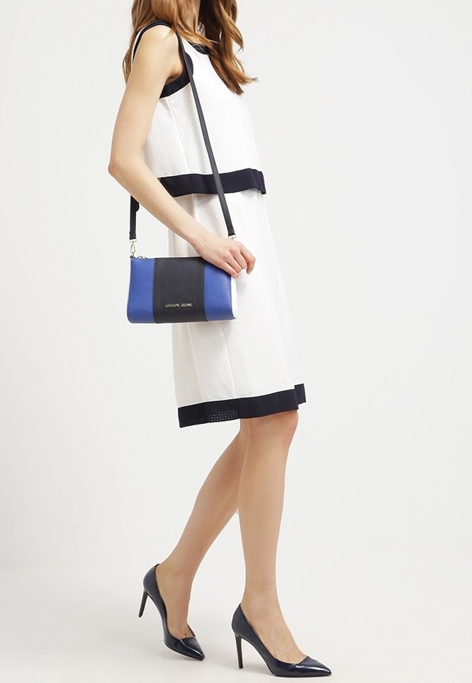 Bílé šaty s námornickými horizontálními pruhy, doplněné modro-černou kabelkou přes rameno. Modelka má na sobě námornické vysoké podpatky. Hladká textura.