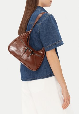 Frau trägt ein kurzärmeliges Hemd aus blauer Jeans und weiße Hose, sie trägt eine braune Leder-Umhängetasche mit Schnallen- und Reißverschlussdetails.