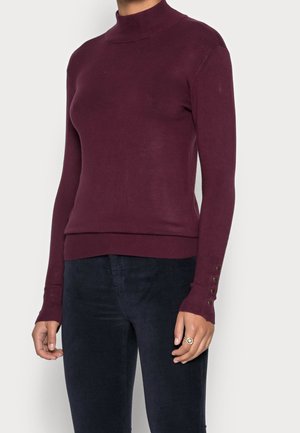 Personne portant un pull à col roulé bordeaux ajusté à manches longues avec des détails de boutons aux poignets et un pantalon sombre, debout devant un fond uni.