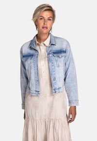 Hellblaues Jeansjacke mit verwaschener Vorderseite, zwei Brusttaschen und silbernen Knöpfen; getragen über einem cremefarbenen gemusterten Kleid.