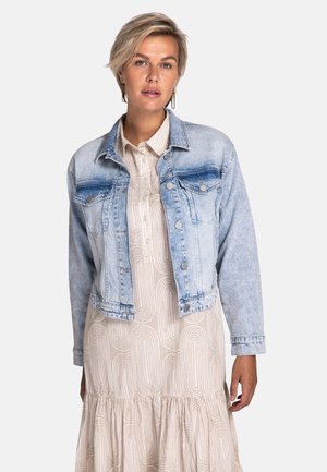 Hellblaues Jeansjacke mit verwaschener Vorderseite, zwei Brusttaschen und silbernen Knöpfen; getragen über einem cremefarbenen gemusterten Kleid.