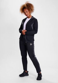 Svart sportjacka med grå detaljer och dragkedja, kombinerad med svarta byxor med fiskbensrand. Modellen har på sig svarta sportskor.