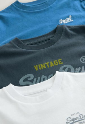 Három kerek nyakú póló felhalmozva: kék, sötétszürke és fehér. Pamut anyag, szitázott "SuperDry" logók, és "VINTAGE" sárgában a szürke pólón.