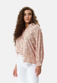 Light pink button-up shirt with white polka dots, long sleeves, and a relaxed fit. Paired with white pants, showcasing a casual look.