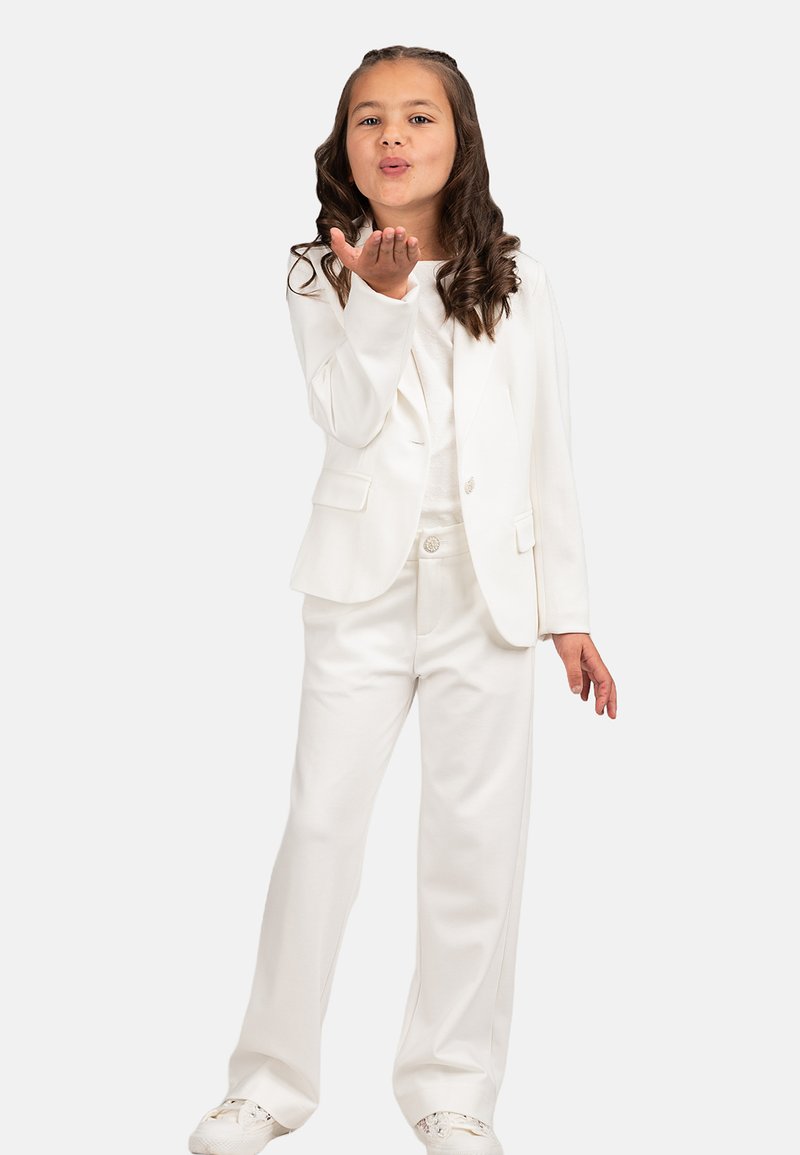 Weißer, maßgeschneiderter Anzug mit tailliertem Sakko und weit geschnittenen Hosen. Das Mädchen macht mit einer Hand spielerische Gesten, ihre lockigen Haare fallen locker herunter.