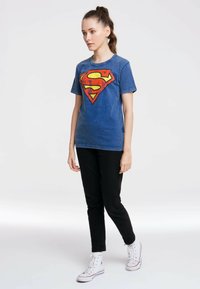 Denim t-shirt in blue featuring a large red and yellow Superman logo, short sleeves, and a round neckline, paired with black pants and white sneakers.