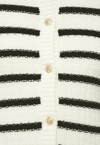 Λευκή πλεκτή ζακέτα με μαύρες οριζόντιες ρίγες, χρυσά κουμπιά με σχέδιο άγκυρας και ριμπ πλεκτή υφή κατά μήκος των άκρων.