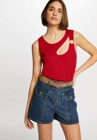 Rotes ärmelloses Top mit einem Ausschnitt-Design und einem goldenen Ring-Akzent, kombiniert mit blauen Jeansshorts mit vorderen Taschen und einem khakifarbenen Gürtel.