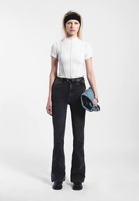 Haut blanc ajusté avec couture verticale, jean noir évasé, et une veste en denim bleu tenue à la main. Accessoire serre-tête noir.