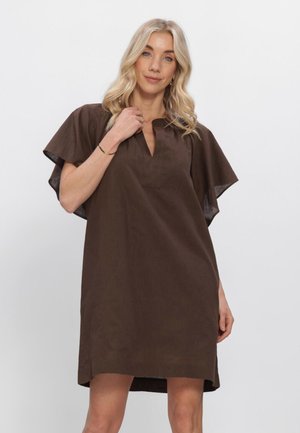 Femme blonde aux cheveux ondulés portant une robe marron ample à larges manches, debout devant un fond uni gris clair.