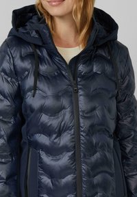 Femme portant une veste à capuche matelassée bleu marine avec une fermeture éclair sur le devant et des cordons noirs par-dessus une chemise crème.