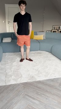 Zwarte V-hals t-shirt, oranje gestreepte shorts, bruine boot schoenen. Staande op een wit tapijt, in een kamer met een blauwe sectionele bank en kussens.