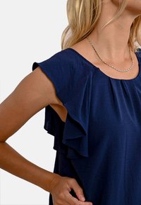 Top blu navy con maniche a volant, realizzato in tessuto morbido, presenta un collo a giro e una vestibilità comoda. Collo adornato con una delicata catena.