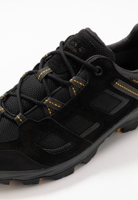 Scarpe da ginnastica nere realizzate con materiali in rete e camoscio, con dettagli di cuciture gialle, un colletto imbottito e una suola nera robusta.