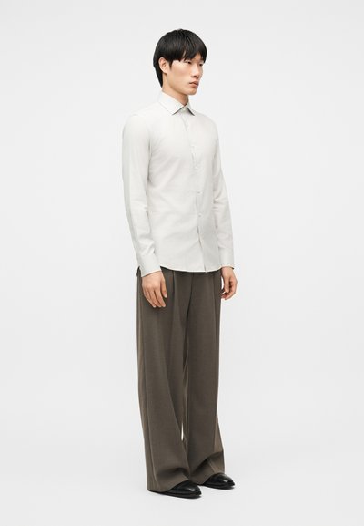 Άνδρας που στέκεται, φοράει εφαρμοστό λευκό πουκάμισο με μακριά μανίκια και κουμπιά, άνετο καφέ παντελόνι και μαύρα παπούτσια μπροστά σε απλό φόντο.