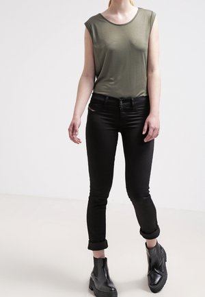 Vrouw die een olijfgroene mouwloze top draagt, zwarte opgerolde skinny jeans en zwarte enkellaarzen, staand tegen een effen witte achtergrond.