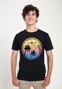 Sort t-shirt med et grafisk design, der inkluderer palmer, et fly og farverige solnedgangsgradienter; rund logo-tekst "Pan American Flight Club."