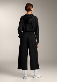 Zwarte hoodie met een relaxed fit en trekkoordcapuchon, gecombineerd met wijde zwarte broek. Geaccentueerd met witte sokken en lichtgrijze sneakers.