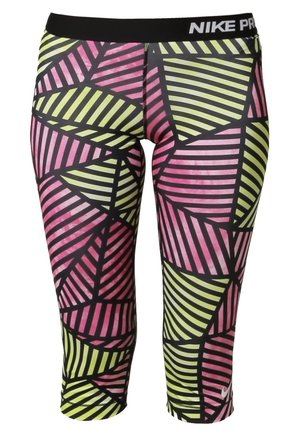 Kobieta ubrana w biały top, wielokolorowe geometryczne legginsy capri i neonowo żółte z białymi butami sportowymi, idąca z lekko opuszczoną głową.