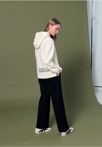 Kremowa bluza z kapturem z logo na plecach, zestawiona z luźnymi czarnymi spodniami i białymi sneakersi z czarnymi akcentami. Tło studyjne w zielonym kolorze.