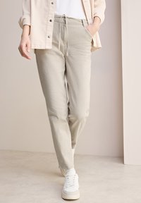 Beżowe spodnie z korduroyu z elastycznym pasem, bocznymi kieszeniami i zwężanym krojem. Zestawione z białymi tenisówkami na jasnym tle.