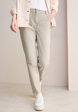 Pantalons en velours côtelé beige avec une taille à cordon de serrage, des poches latérales et une coupe fuselée. Associés à des baskets blanches sur un fond clair.