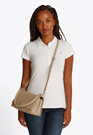 Ženska z pletenicami, oblečena v belo polo majico in temne kavbojke, nosi bež usnjeno torbico čez ramo s srebrno okovje.