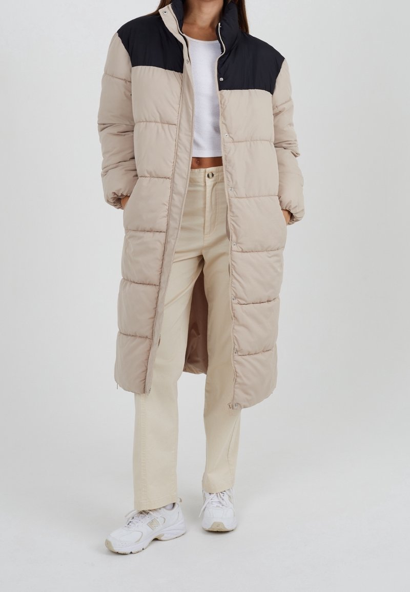 Abrigo acolchado con cuerpo beige y panel superior negro, botones a presión y bolsillos laterales. Se lleva con pantalones de color claro y zapatillas.