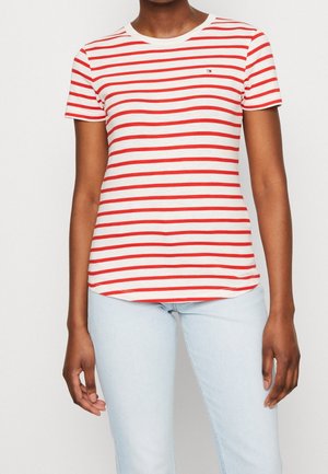 Persoon draagt een wit shirt met korte mouwen en horizontale rode strepen en lichtblauwe spijkerbroek, staand tegen een effen achtergrond.