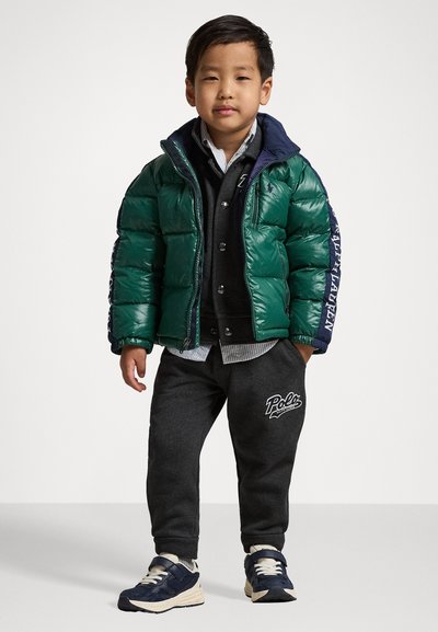Groene pufferjack met marineblauwe accenten, grijze sweatshirt en donkergrijze joggers. Witte logo op de broek en blauwe sneakers met lichte zolen.