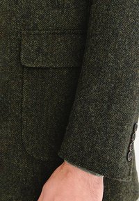 Marynarka tweedowa w ciemnozielonym kolorze z wzorem jodełki, wyposażona w kieszeń na piersi i mankiety z guzikami. Teksturowana tkanina wełniana.