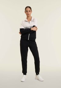 Donna giovane in piedi con le braccia incrociate, indossa una giacca zip bianca e nera, pantaloni jogging neri e sneakers bianche su sfondo semplice.