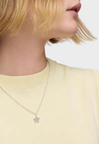Collana d'argento con un pendente a forma di fiore, indossata sopra un maglione a coste di un giallo chiaro, con un primo piano del colletto e i dettagli della gioielleria.