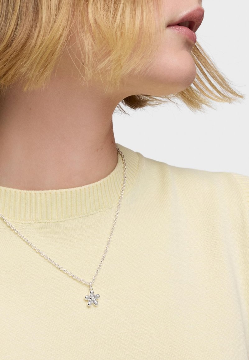 Collana d'argento con un pendente a forma di fiore, indossata sopra un maglione a coste di un giallo chiaro, con un primo piano del colletto e i dettagli della gioielleria.