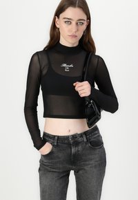 Black, long-sleeve, sheer crop top with mock neck; features white text "Thanks OK Bye." Accompanied by grey jeans and a black bag.