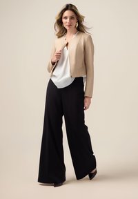 Veste en simili cuir beige avec un col rond, associée à un chemisier blanc et un pantalon large noir. Accessoirisée avec des escarpins noirs.