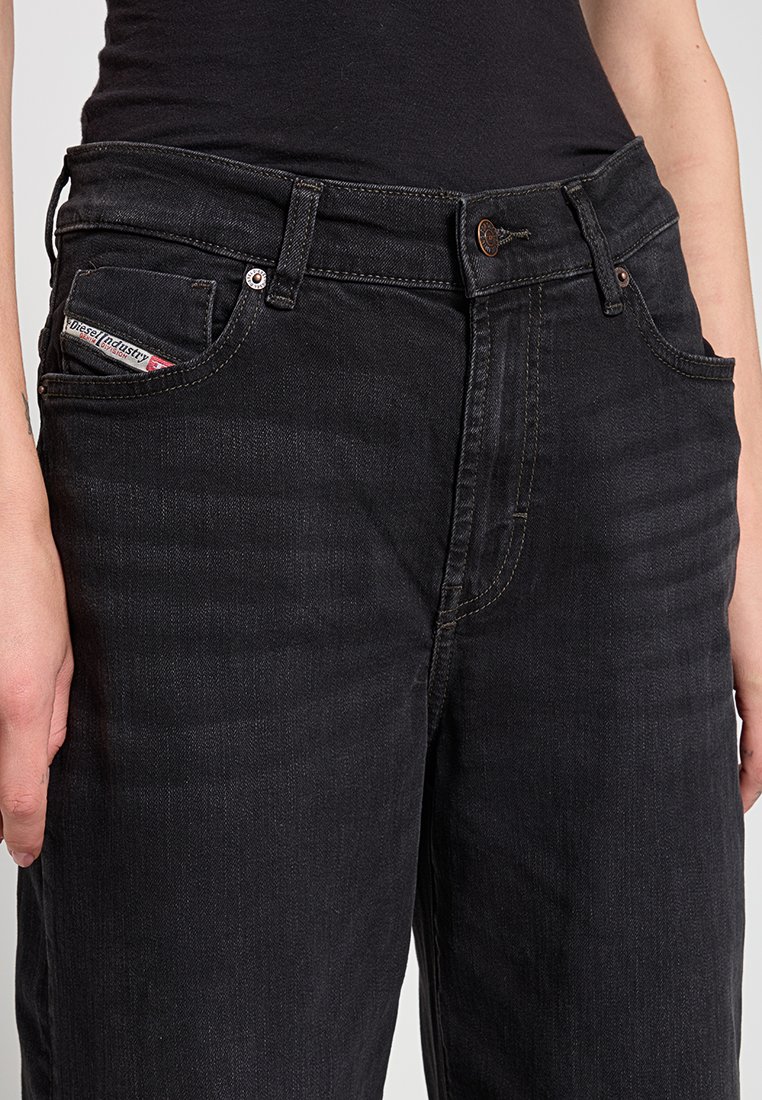 Nahaufnahme einer Person, die hochgeschnittene schwarze Jeans und ein schwarzes Oberteil trägt. Die Vorderentaschen und das Diesel Industry-Label auf der kleinen Tasche sind zu sehen.