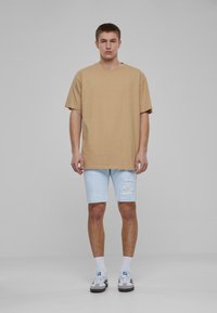 Camiseta de algodón de color marrón claro, de gran tamaño y con mangas cortas, combinada con unos pantalones cortos de mezclilla azul claro con efecto desgastado. El modelo lleva puestos calcetines blancos y zapatillas grises.