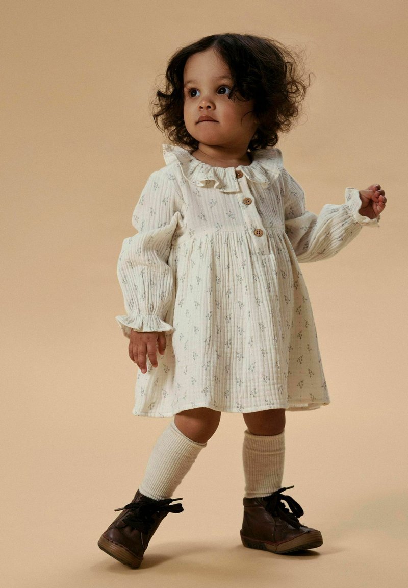 Bambino con capelli ricci che indossa un vestito crema a fiori, calzini fino al ginocchio e stivali stringati marrone scuro su uno sfondo beige.