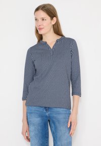Femme portant une chemise à manches longues rayée verticalement bleu et blanc, et un jean bleu clair, regardant légèrement vers sa gauche.