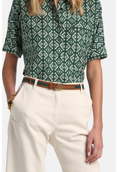 Femeie purtând o cămașă cu mânecă scurtă, cu model verde, băgată în pantaloni crem, cu o curea verde și maro și o brățară cu lanț auriu.