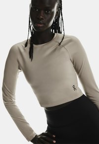 Crop top beige à manches longues en matériau lisse, avec un col arrondi et un logo noir imprimé sur le côté gauche. Pantalons noirs taille haute.
