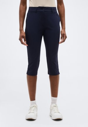 Άτομο που φοράει στενά παντελόνια σε σκούρο μπλε χρώμα, συνδυασμένα με λευκά αθλητικά παπούτσια και λευκές κάλτσες αστραγάλου, μπροστά σε απλό φόντο.